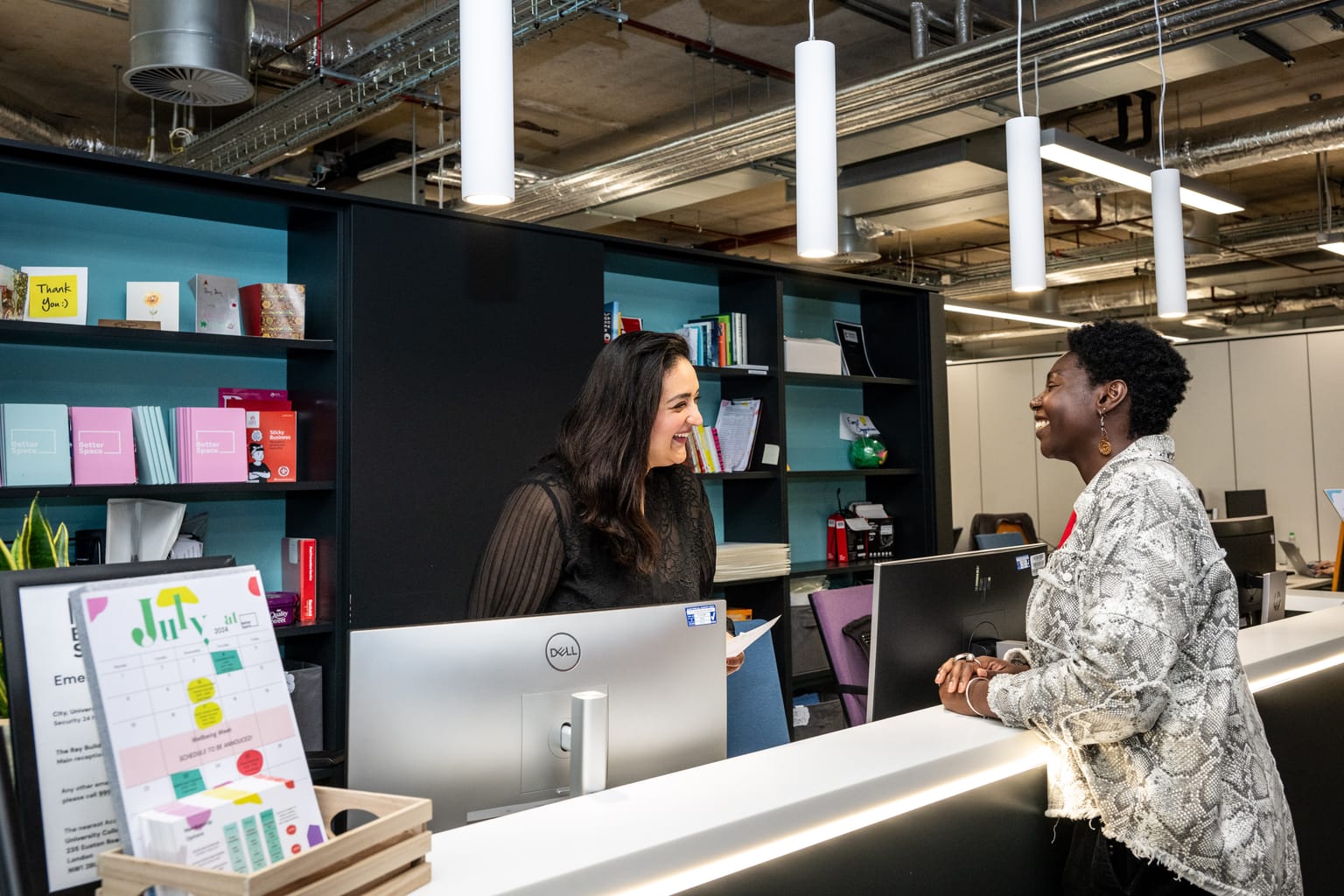 Two women are interacting and smiling at a reception desk in a modern office setting, showcasing the vibrant atmosphere of startup success. The background features shelves with books and office supplies. The desk has a computer monitor, papers, and a colorful calendar. Overhead lights and an industrial ceiling are visible.